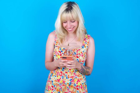 Téléchargez les photos : Sourire fille caucasienne portant robe florale isolé sur fond bleu en utilisant un téléphone portable, messagerie, être heureux de texter avec des amis, en regardant smartphone. Technologies modernes et communication. - en image libre de droit