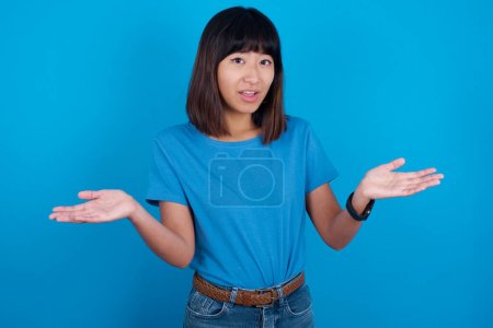 Téléchargez les photos : Et alors ? Portrait d'une jeune femme asiatique arrogante portant un t-shirt bleu sur fond bleu haussant les mains latéralement souriant haletant indifférent, disant quelque chose d'évident. - en image libre de droit