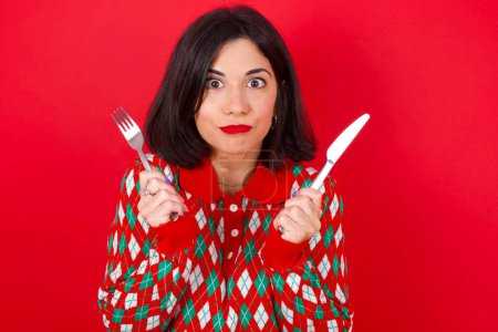 Téléchargez les photos : Affamé brunette femme caucasienne portant chandail de Noël sur fond rouge tenant dans la main couteau fourchette veulent délicieuse pizza tarte - en image libre de droit