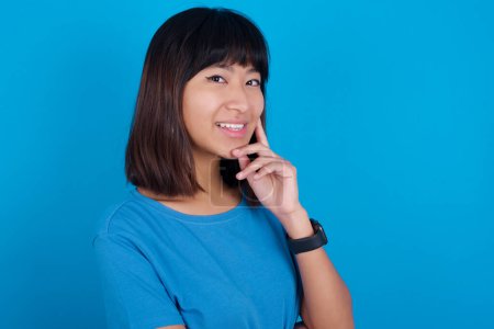 Téléchargez les photos : Insouciant succès jeune femme asiatique portant t-shirt bleu sur fond bleu touchant mâchoire regardant caméra inclinaison tête souriant dents blanches ravi. Concept de soins dentaires. - en image libre de droit