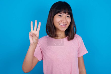 Téléchargez les photos : Jeune femme asiatique portant rose t-shirt sur fond bleu montrant et pointant vers le haut avec les doigts numéro trois tout en souriant confiant et heureux. - en image libre de droit