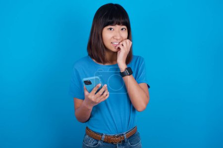 Foto de Miedo divertido joven asiático mujer usando azul camiseta contra fondo azul celebración de teléfono y morder las uñas - Imagen libre de derechos