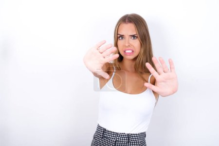 Dissatisfied Young Caucasian girl wearing white tank top on white background frowns face, has disgusting expression, shows tongue, expresses non compliance, irritated with somebody.