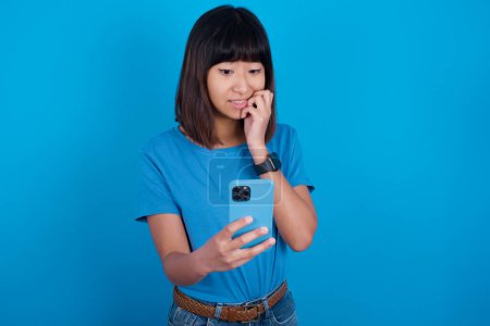 Foto de Retrato de una joven asiática bastante asustada usando una camiseta azul contra un fondo azul charlando con uñas mordidas después de leer algunas noticias aterradoras en su teléfono inteligente. - Imagen libre de derechos