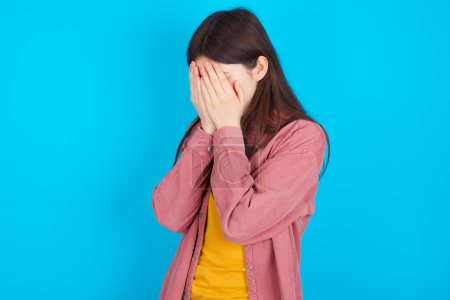 Photo for Sad young caucasian girl wearing pink shirt isolated over blue background covering face with hands and crying. - Royalty Free Image