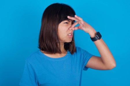 Téléchargez les photos : Mécontent jeune femme asiatique portant t-shirt bleu sur fond bleu bouchons nez comme sent quelque chose puant et désagréable, se sent aversion, déteste parfum dégoûtant. - en image libre de droit