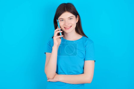 Téléchargez les photos : Portrait d'une jeune caucasienne souriante portant un t-shirt bleu isolé sur fond de studio bleu parlant sur téléphone portable. Concept d'entreprise, confiance et communication. - en image libre de droit