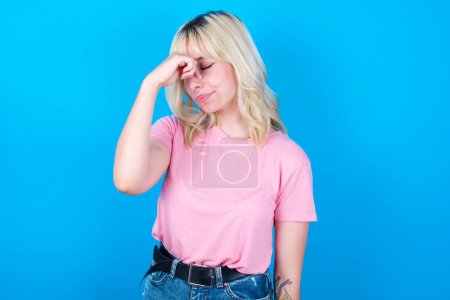 Téléchargez les photos : Fille caucasienne très contrariée portant t-shirt rose isolé sur fond bleu touchant le nez entre les yeux fermés, veut pleurer, ayant une relation stressante ou ayant des problèmes avec le travail - en image libre de droit
