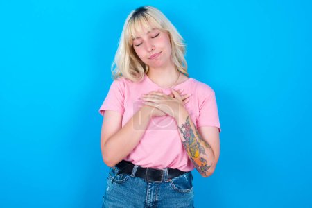 Photo for Caucasian girl wearing pink t-shirt isolated over blue background closes eyes and keeps hands on chest near heart, expresses sincere emotions, being kind hearted and honest. Body language and real feelings concept. - Royalty Free Image