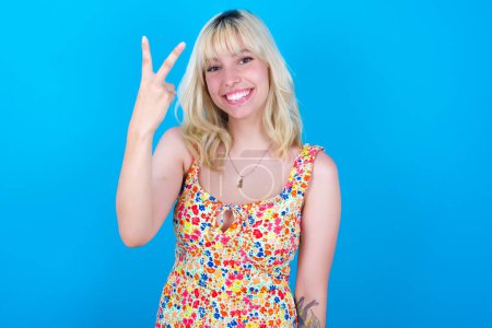 Photo for Caucasian girl wearing floral dress isolated over blue background smiling and looking friendly, showing number two or second with hand forward, counting down - Royalty Free Image