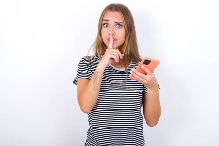 beautiful blonde girl wearing striped t-shirt on white background holding modern gadget ask not tell secrets