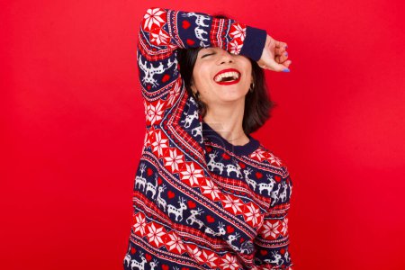 Foto de Morena mujer caucásica vistiendo suéter de Navidad sobre fondo rojo cubriendo los ojos con el brazo sonriendo alegre y divertido. Concepto ciego. - Imagen libre de derechos