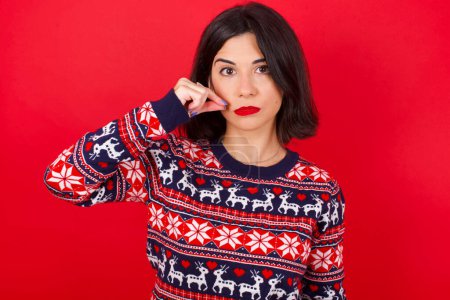 Foto de Morena mujer caucásica vistiendo suéter de Navidad sobre fondo rojo boca y labios cerrados como cremallera con los dedos. Secreto y silencioso, tabú hablando. - Imagen libre de derechos