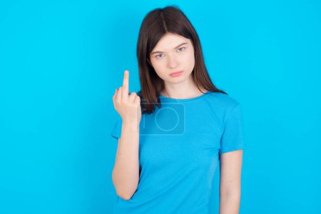 Foto de Joven chica caucásica con camiseta azul aislado sobre fondo estudio azul muestra dedo medio mala señal pide no molestarse. Provocación y actitud grosera. - Imagen libre de derechos