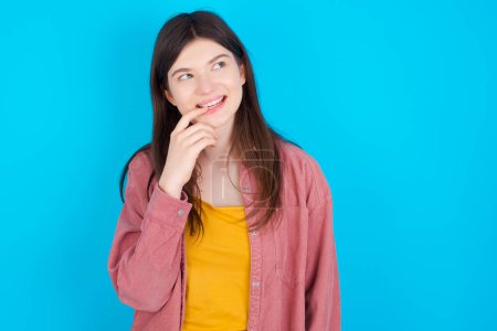 Photo for Young caucasian girl wearing pink shirt isolated over blue background with thoughtful expression, looks to the camera, keeps hand near face, biting a finger thinks about something pleasant. - Royalty Free Image