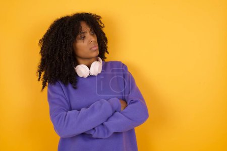 Téléchargez les photos : Portrait de jeune femme afro-américaine expressive avec écouteurs sur fond jaune avec bras croisés - en image libre de droit