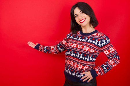 Téléchargez les photos : Brune caucasienne portant un pull de Noël sur fond rouge se sentant heureux et joyeux, souriant et vous accueillant, vous invitant à entrer avec un geste amical - en image libre de droit