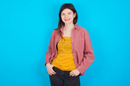 Téléchargez les photos : Portrait de jeune fille caucasienne réussie portant chemise rose isolée sur fond bleu souriant largement avec expression auto-assurée. Confiance et concept d'entreprise. - en image libre de droit