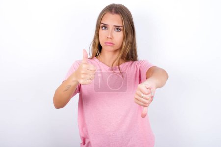 Foto de Joven chica caucásica vistiendo camiseta rosa sobre fondo blanco sintiéndose inseguro haciendo buena mala señal. Descontento y poco impresionado. - Imagen libre de derechos