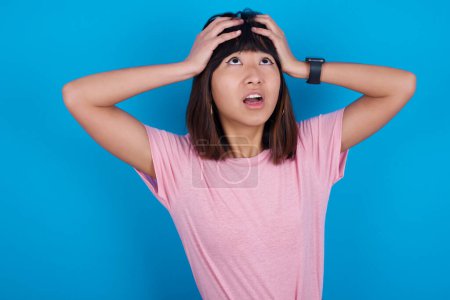 Photo for Horrible, stress, shock. Portrait emotional crazy young asian woman wearing pink t-shirt against blue background clasping head in hands. Emotions, facial expression concept. - Royalty Free Image