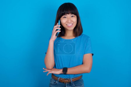Téléchargez les photos : Portrait d'une jeune femme asiatique souriante portant un t-shirt bleu sur fond bleu parlant sur téléphone portable. Concept d'entreprise, confiance et communication. - en image libre de droit