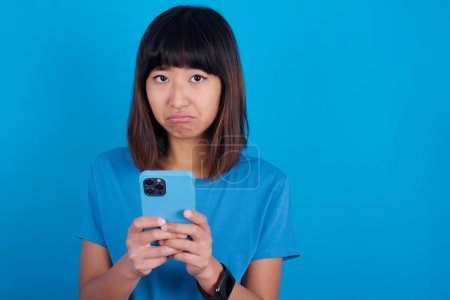 Photo for Portrait of a confused young asian woman wearing blue t-shirt against blue background holding mobile phone and shrugging shoulders and frowning face. - Royalty Free Image