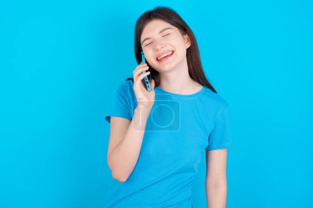 Foto de Divertido joven caucásico chica usando azul camiseta aislado sobre azul fondo del estudio ríe felizmente, tiene conversación telefónica, siendo divertido por amigo, cierra los ojos. - Imagen libre de derechos