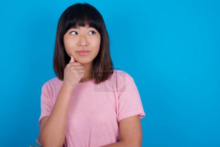 Photo for Portrait of thoughtful young asian woman wearing pink t-shirt against blue background keeps hand under chin, looks away trying to remember something or listens something with interest. Youth concept. - Royalty Free Image
