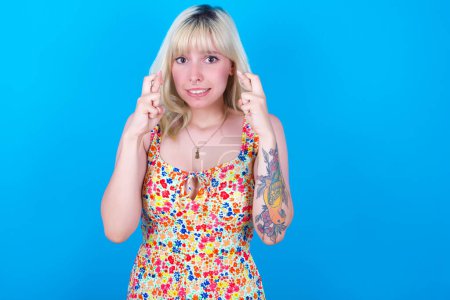 Photo for Caucasian girl wearing floral dress isolated over blue background holding fingers crossed with worried expression hoping boss didn't noticed mistakes at work. - Royalty Free Image