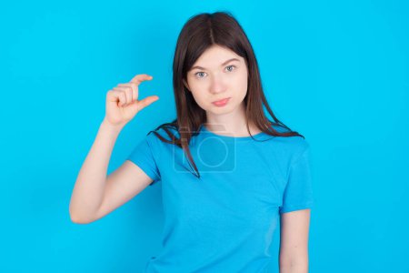 Foto de Joven chica caucásica con camiseta azul aislado sobre azul estudio monederos labio y gestos con la mano, muestra algo muy poco. - Imagen libre de derechos