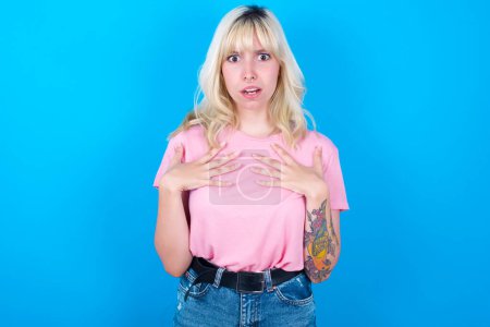 Photo for Caucasian girl wearing pink t-shirt isolated over blue background keeps hands on chest feeling shocked and scared, mouth widely opened, stares at camera saying: Who, me? - Royalty Free Image