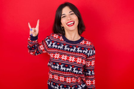 Foto de Morena mujer caucásica vistiendo jersey navideño sobre fondo rojo haciendo un gesto de roca y sonriendo a la cámara. Listo para ir a su concierto favorito de la banda. - Imagen libre de derechos