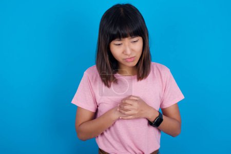 Photo for Sad young asian woman wearing pink t-shirt against blue background feeling upset while spending time at home alone staring at camera with unhappy or regretful look. - Royalty Free Image