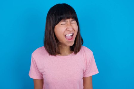 Photo for Stressful young asian woman wearing pink t-shirt against blue background screams in panic, closes eyes in terror, keeps hands on head, finds out terrified news, can't believe it. - Royalty Free Image