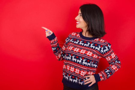 Photo for Smiling brunette caucasian woman wearing christmas sweater over red background indicating finger empty space showing best low prices - Royalty Free Image