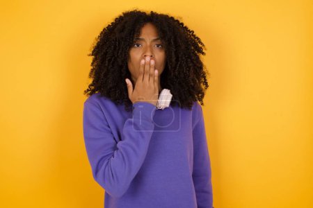 Téléchargez les photos : Portrait de jeune femme afro-américaine expressive avec écouteurs sur fond jaune couvrant sa bouche - en image libre de droit