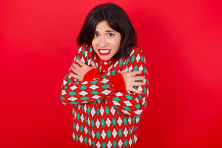 Photo for Desperate brunette caucasian woman wearing christmas sweater over red background trembles and feels cold, hugs oneself to warm up or feels scared notices something terrifying. - Royalty Free Image