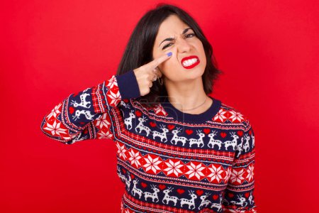 Téléchargez les photos : Femme caucasienne brune portant chandail de Noël sur fond rouge pointant malheureux de boutons sur le front, infection laide de la tête noire. Acné et problème de peau - en image libre de droit