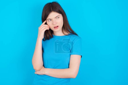 Foto de Sorprendida joven chica caucásica con camiseta azul aislada sobre fondo de estudio azul se ve a un lado sorprendentemente con la boca abierta. - Imagen libre de derechos