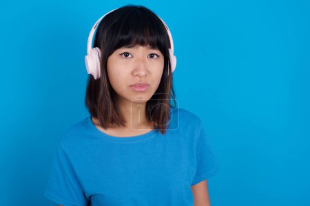 Téléchargez les photos : Grave mécontent jeune femme asiatique portant t-shirt bleu sur fond bleu semble perplexe à la caméra étant en colère porte casque stéréo écoute de la musique tout en marchant dans la rue - en image libre de droit