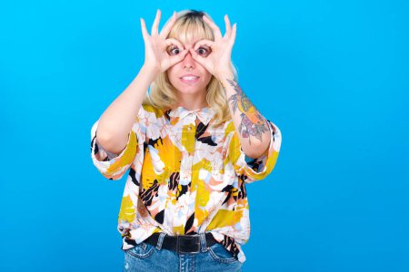Foto de Chica caucásica juguetona emocionada usando camisa floral aislada sobre fondo azul mostrando signo Ok con ambas manos en los ojos, fingiendo usar gafas. - Imagen libre de derechos