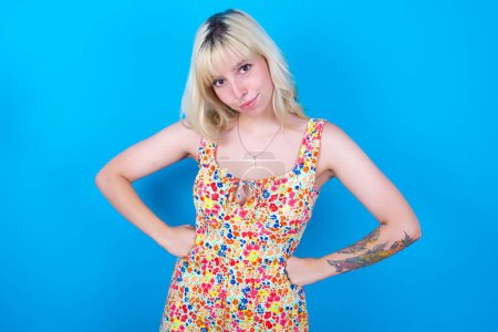 Photo for Funny frustrated caucasian girl wearing floral dress isolated over blue background holding hands on waist and silly looking at awkward situation. - Royalty Free Image