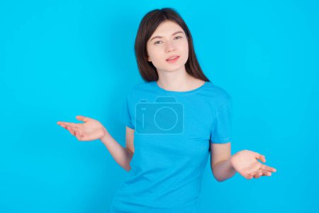 Téléchargez les photos : Jeune fille caucasienne portant bleu t-shirt isolé sur fond bleu studio semble épaules hausseurs d'épaules incertaines. - en image libre de droit