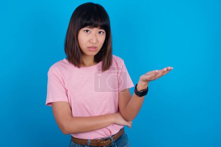 Téléchargez les photos : Studio shot of frustrated young asian woman wearing pink t-shirt against blue background gesturing with raised palm, frowning, being displeased and confused with dumb question. - en image libre de droit