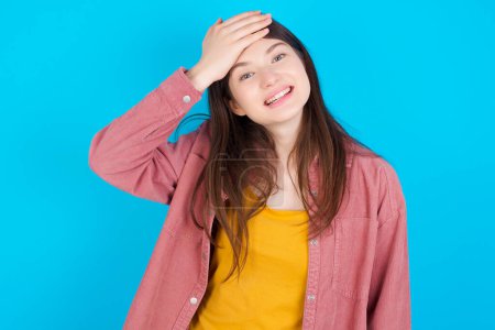 Photo for Oops, what did I do? Young caucasian girl wearing pink shirt isolated over blue background holding hand on forehead with frightened and regret expression. - Royalty Free Image