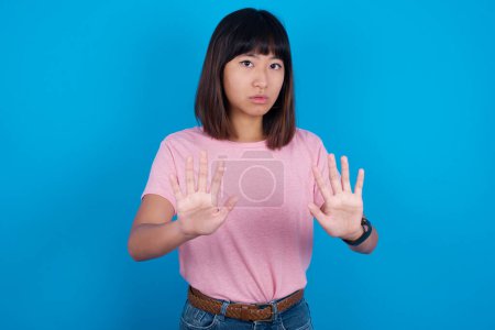 Photo for Young asian woman wearing pink t-shirt against blue background doing stop sing with palm of the hand. Warning expression with negative and serious gesture on the face. - Royalty Free Image