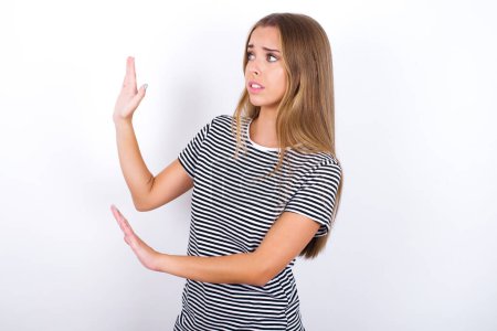 Photo for Displeased beautiful blonde girl wearing striped t-shirt on white background keeps hands towards empty space and asks not come closer sees something unpleasant - Royalty Free Image