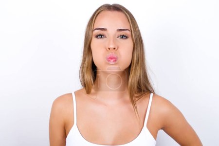 Young Caucasian girl wearing white tank top on white background puffing cheeks with funny face. Mouth inflated with air, crazy expression.