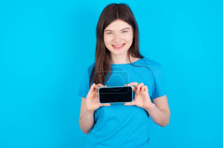 Photo for Cheerful cheery content young caucasian girl wearing blue t-shirt isolated over blue studio background holding in hands device hobby smm post blog - Royalty Free Image