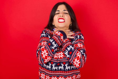 Foto de Morena mujer caucásica vistiendo jersey de navidad sobre fondo rojo gritando sofocado porque estrangulamiento doloroso. Problema de salud. Concepto de asfixia y suicidio. - Imagen libre de derechos